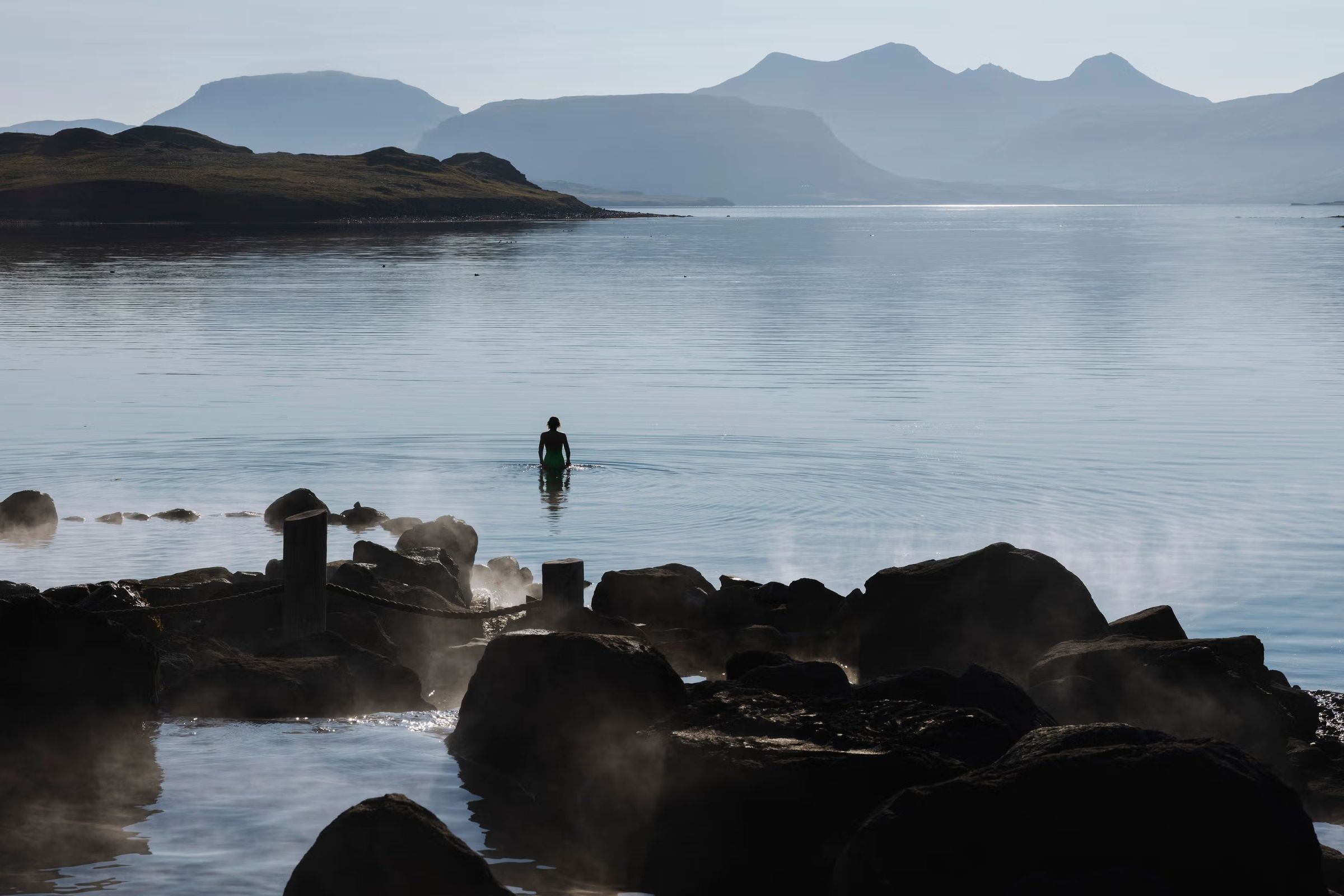 Una persona se adentra en una fuente termal humeante, con rocas oscuras en primer plano y un lago al fondo rodeado de montañas difuminadas por la neblina.