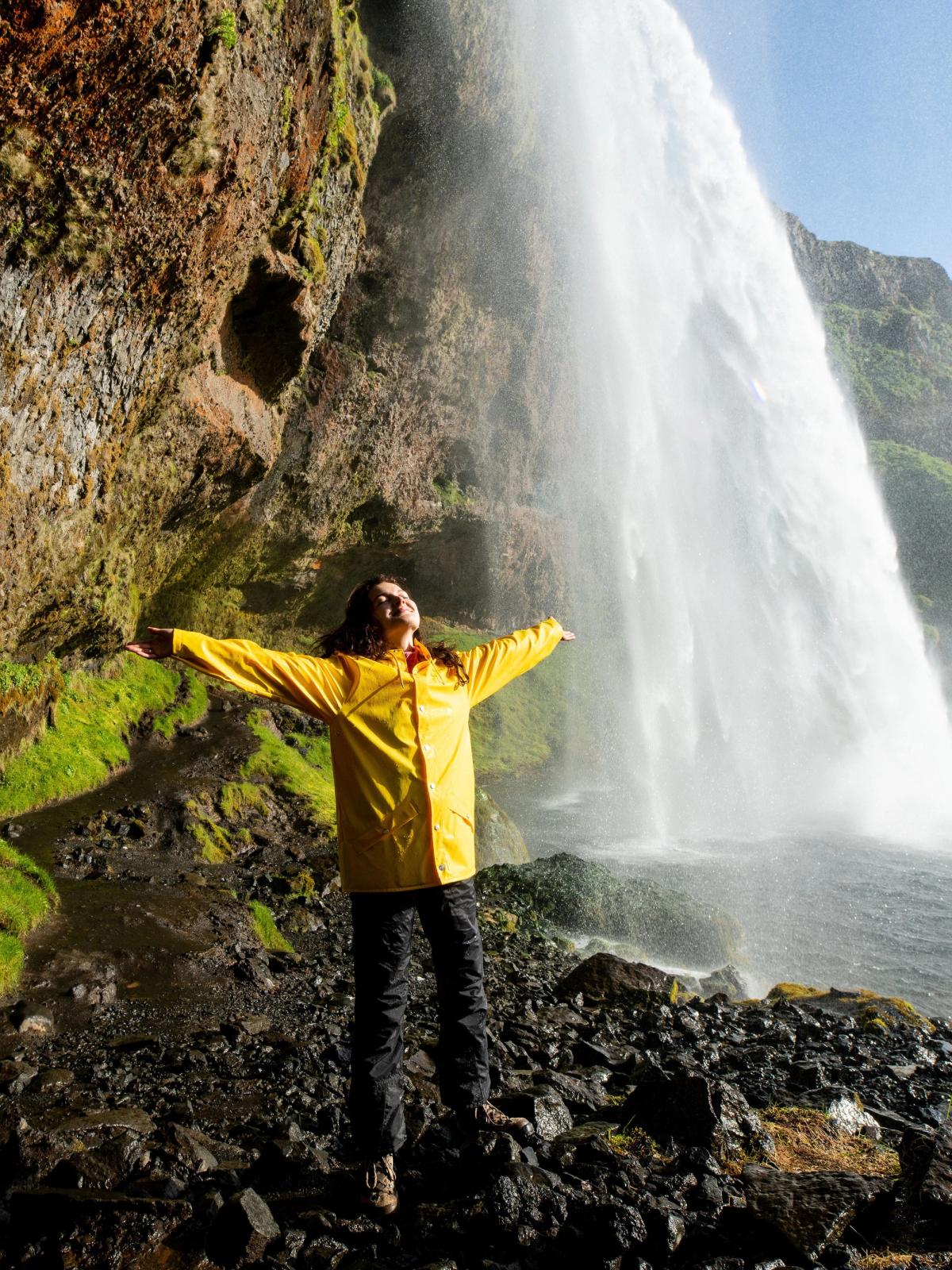 un hombre con un impermeable amarillo está parado frente a una cascada con los brazos extendidos.