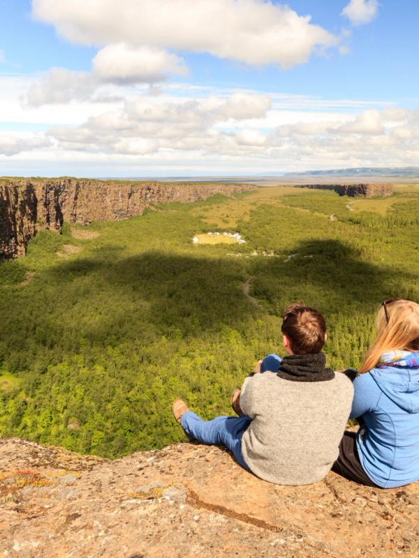 Two people sit on a cliff edge overlooking a wide, tree-filled canyon under a partly cloudy sky.