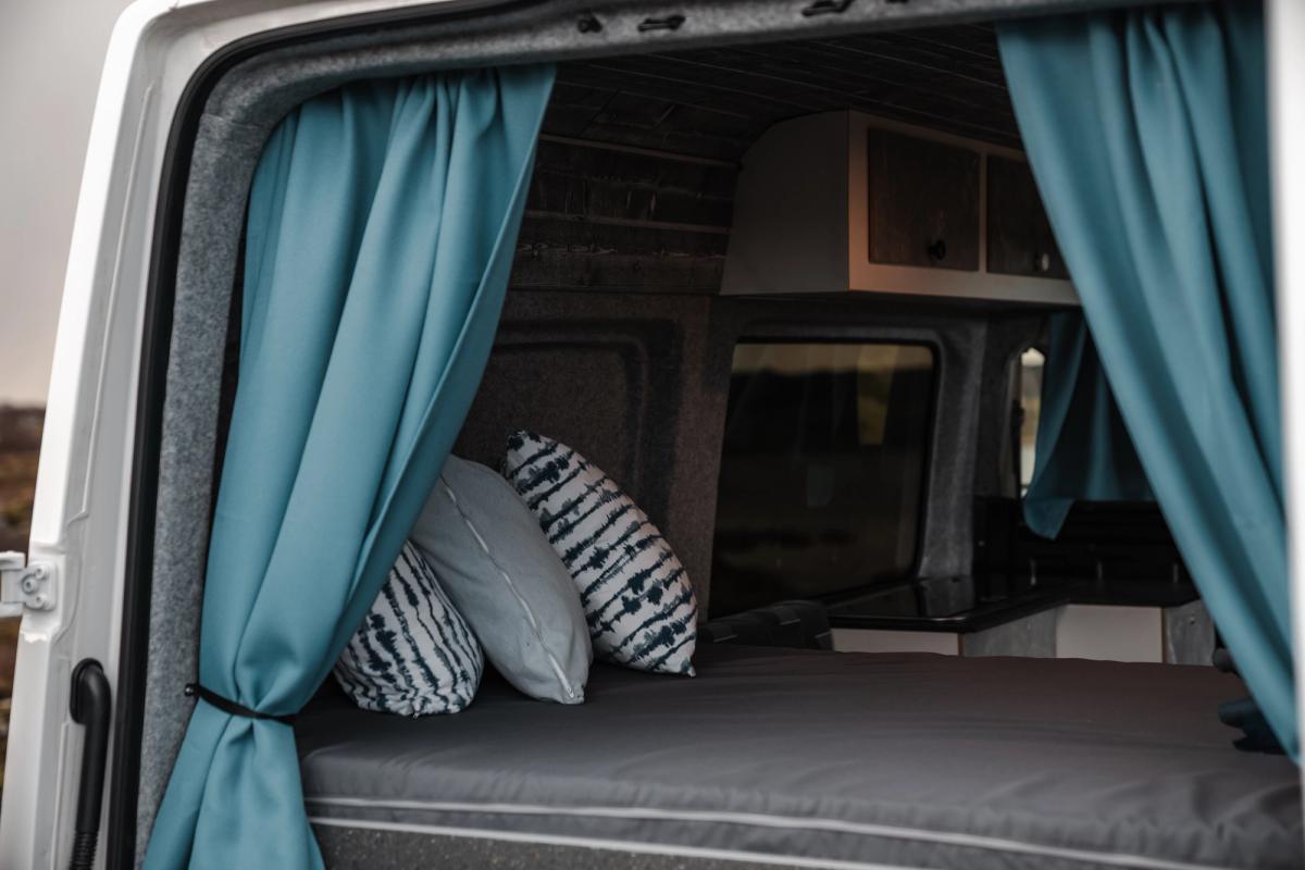 Cama trasera con cojines estampados, enmarcada por cortinas verde azulado y con armarios superiores al fondo
