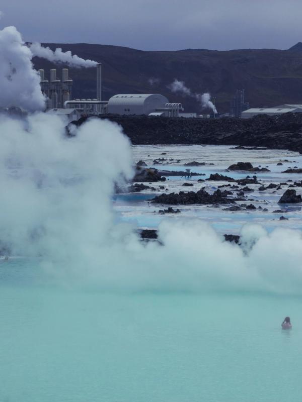Swimmers in a steamy, milky blue geothermal lagoon with a power plant emitting steam in the background.