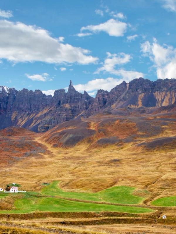 Autumnal landscape with rugged mountains, a spire, fields of green and brown, and white houses under a blue, cloudy sky.