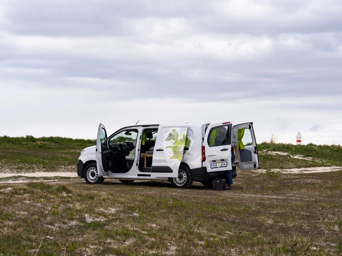 Montaje del campamento con una autocaravana compacta Una autocaravana compacta con capacidad para 2 personas, con las puertas trasera y lateral abiertas, alquiler de autocaravanas en Islandia