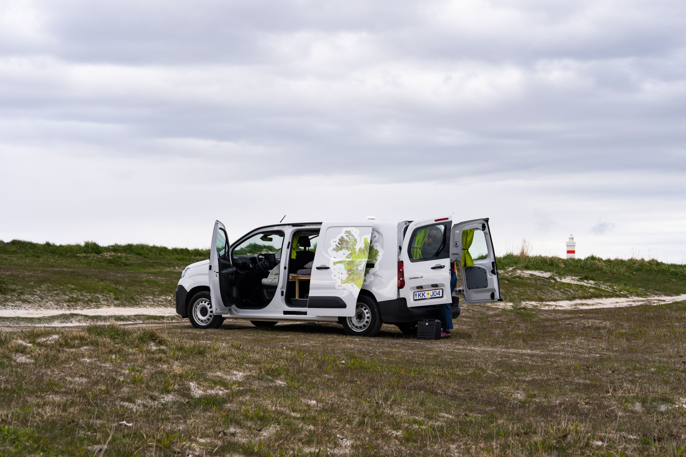 Montaje del campamento con una autocaravana compacta Una autocaravana compacta con capacidad para 2 personas, con las puertas trasera y lateral abiertas, alquiler de autocaravanas en Islandia