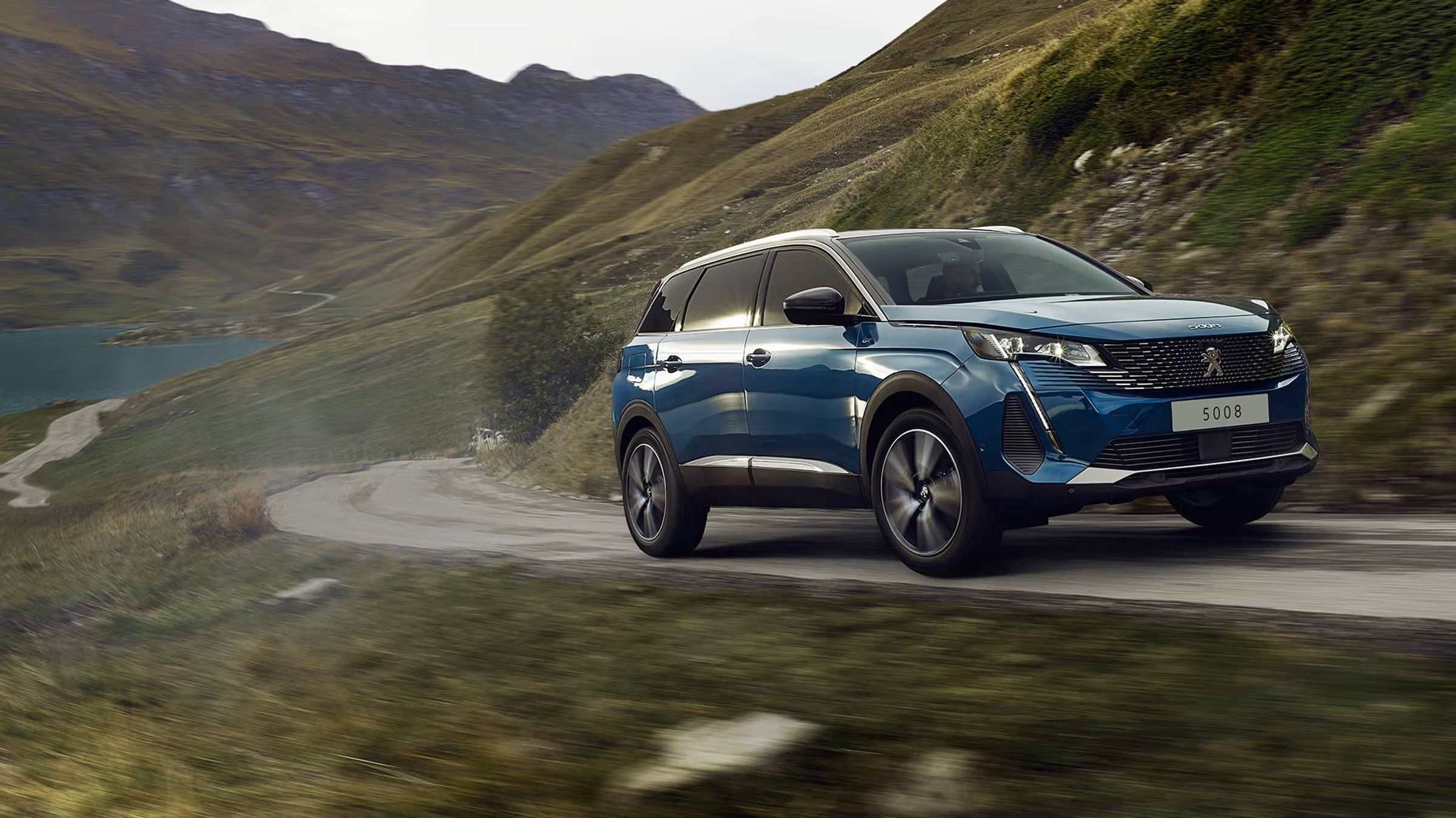 Un Peugeot 5008 SUV azul circula por una carretera de montaña sinuosa junto a un lago.