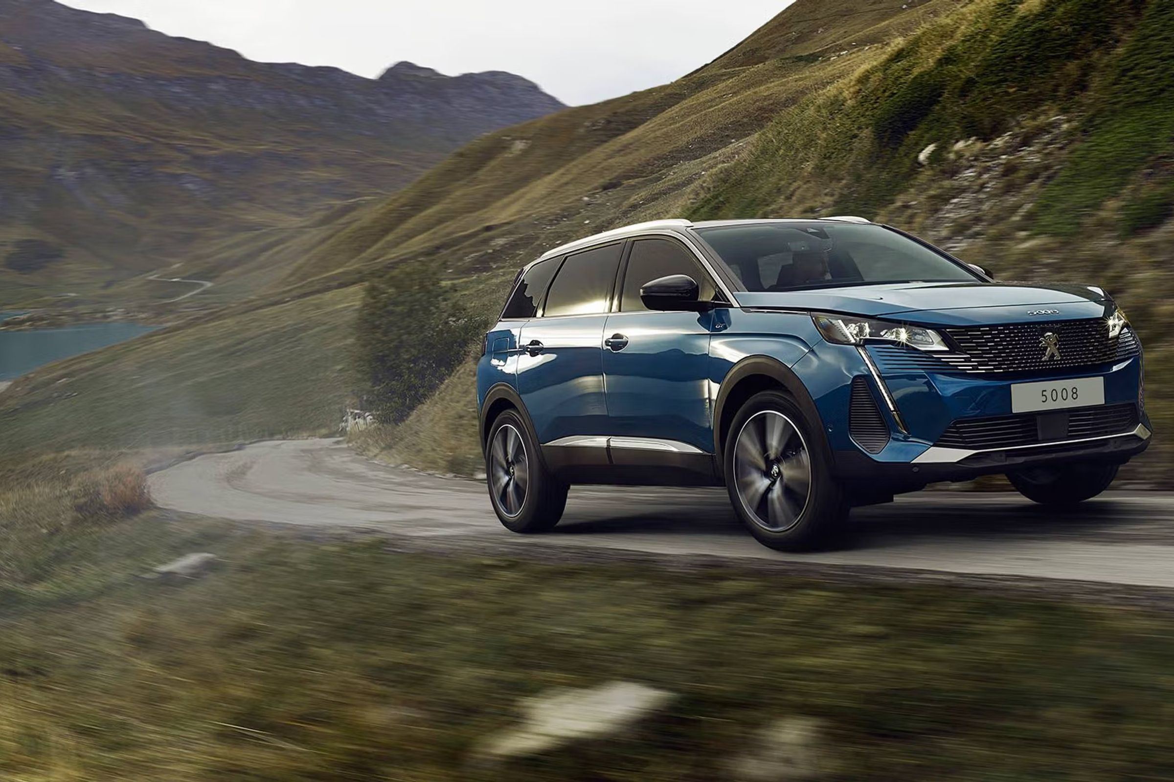Un Peugeot 5008 SUV azul circula por una carretera de montaña sinuosa junto a un lago.