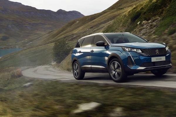 A blue Peugeot 5008 SUV drives on a winding mountain road next to a lake.