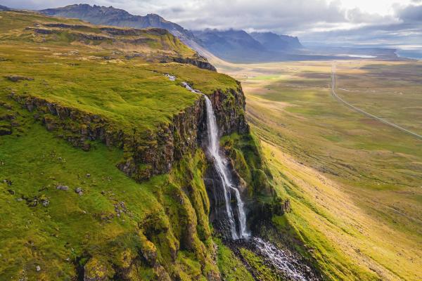 A waterfall cascades down a mossy cliff of vibrant green, overlooking an expansive flat plain with a path and distant mountains under a cloudy sky.
