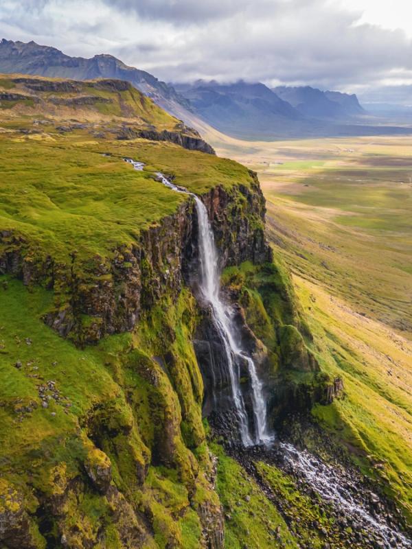 A tall waterfall cascades down a mossy green cliff, overlooking a vast green valley with a winding road and distant mountains under a cloudy sky.