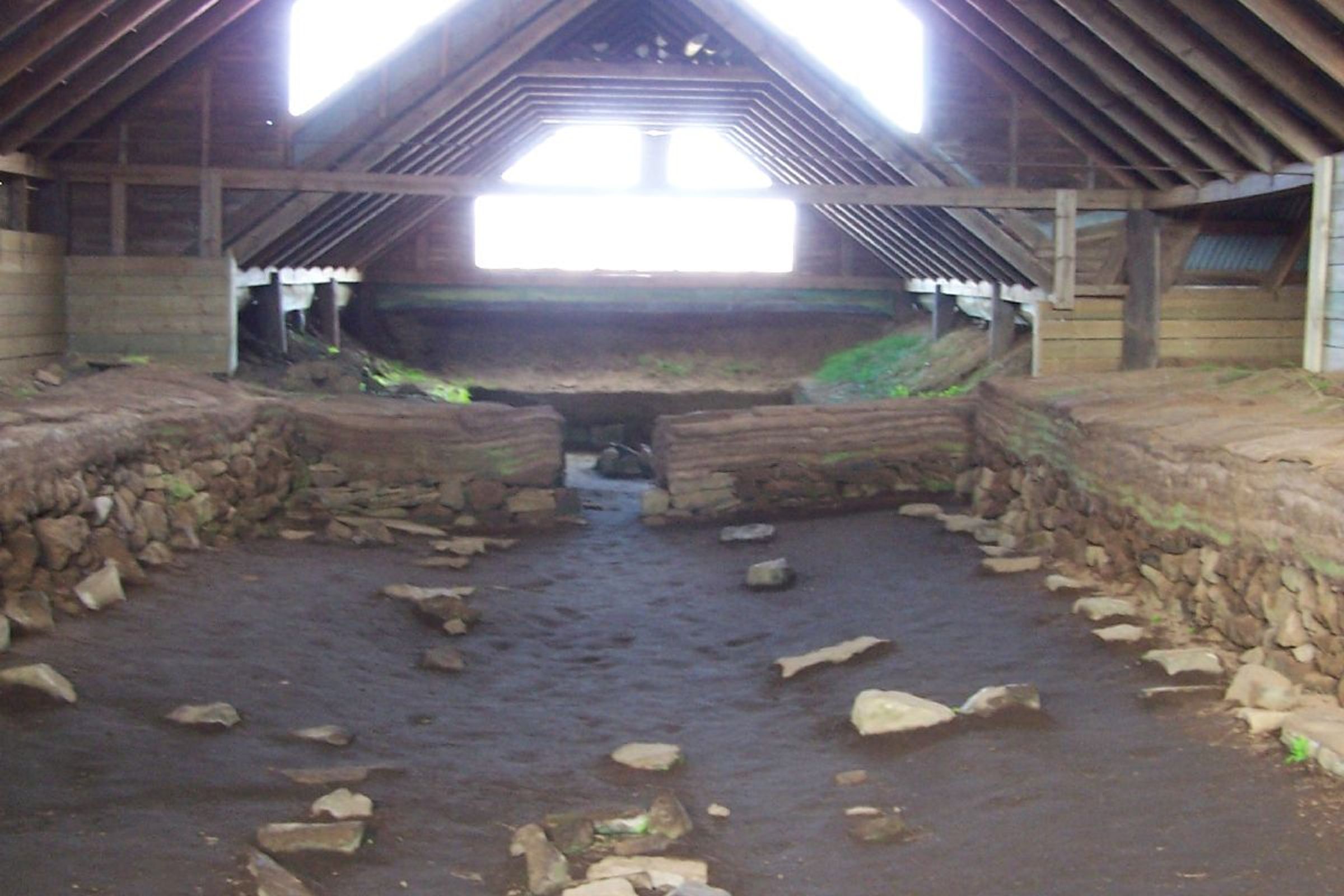 An indoor archaeological site with dark soil, stone foundations, and a high wooden beam roof with bright back windows.