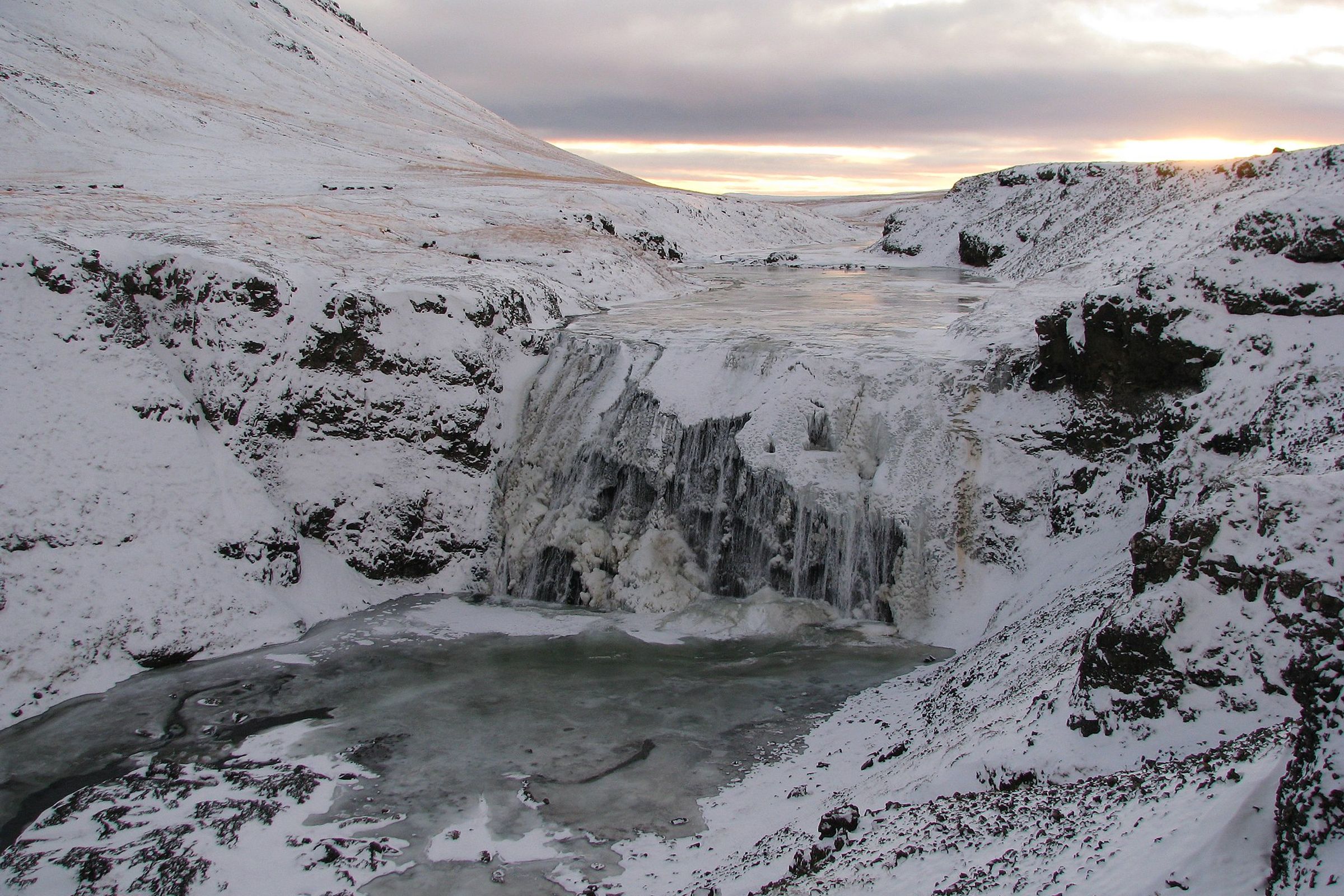 Una cascada parcialmente congelada en un cañón cubierto de nieve bajo un cielo dramático.