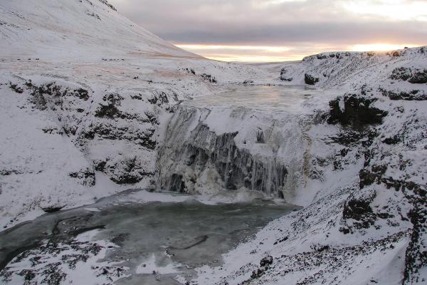 A partially frozen waterfall in a snow-covered canyon under a dramatic sky.