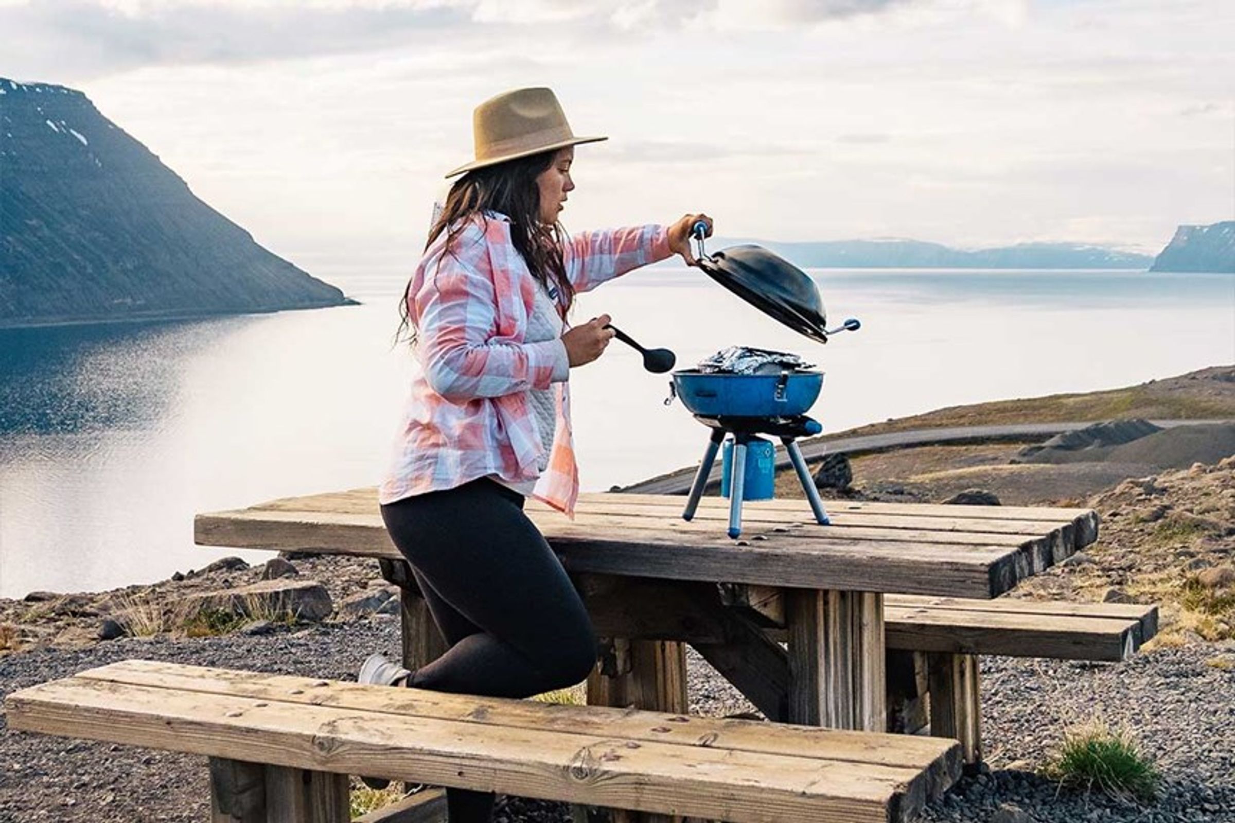 a woman is sitting at a picnic table cooking on a grill in iceland.