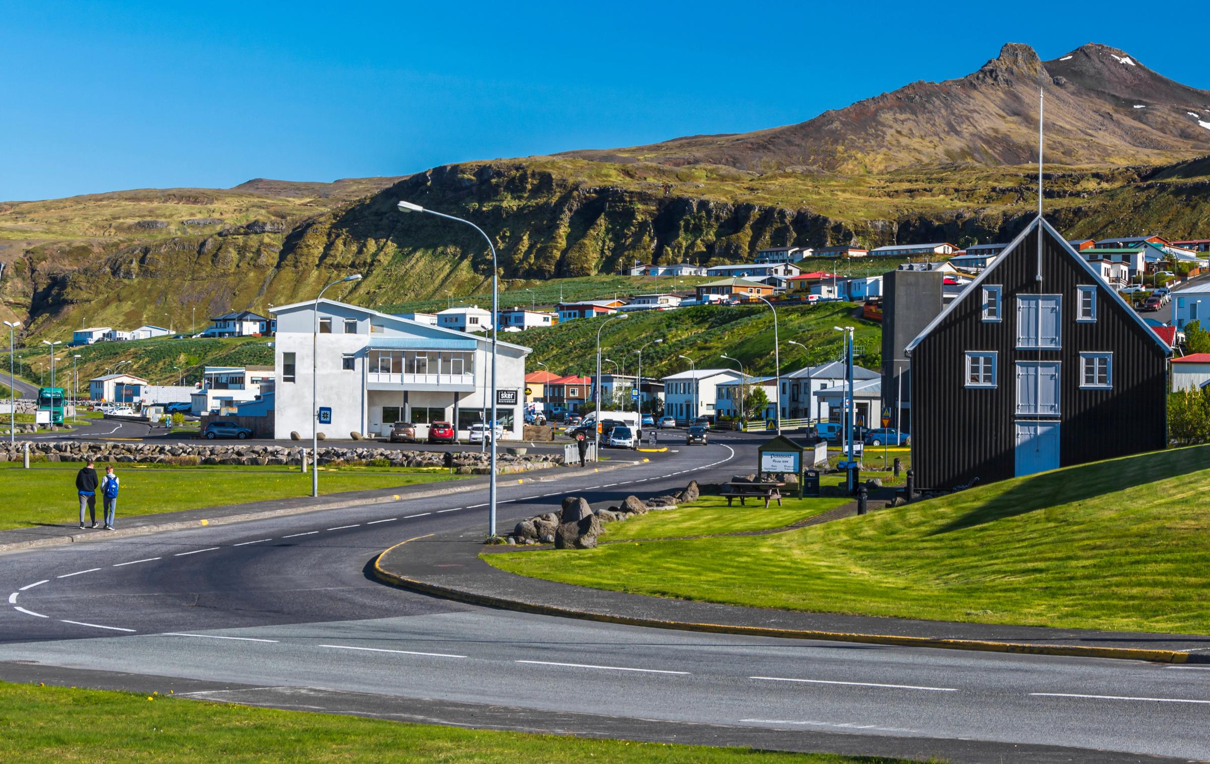 A village with a black wooden house and modern white building on a road, backed by a large green mountain.