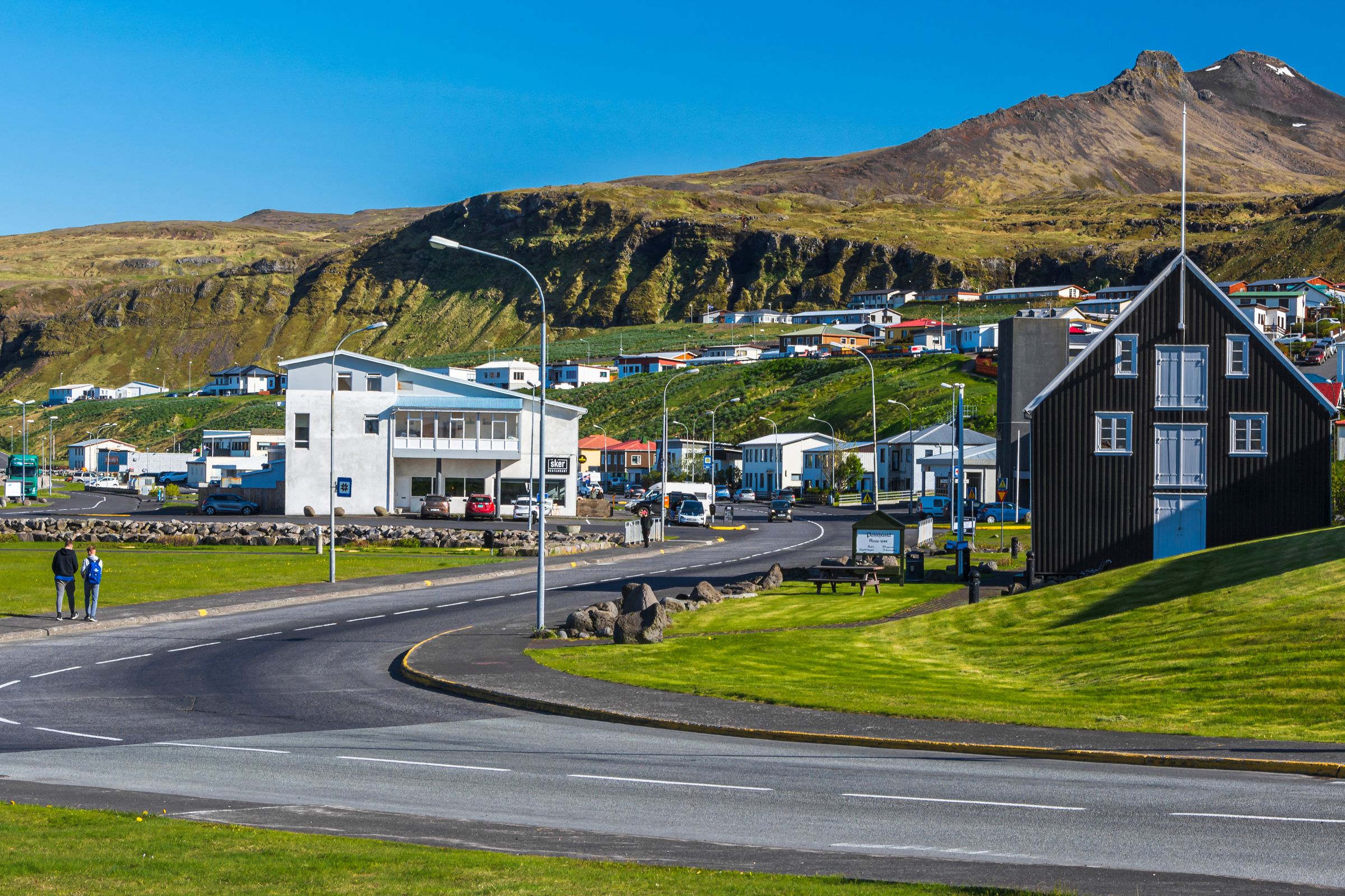 A village with a black wooden house and modern white building on a road, backed by a large green mountain.