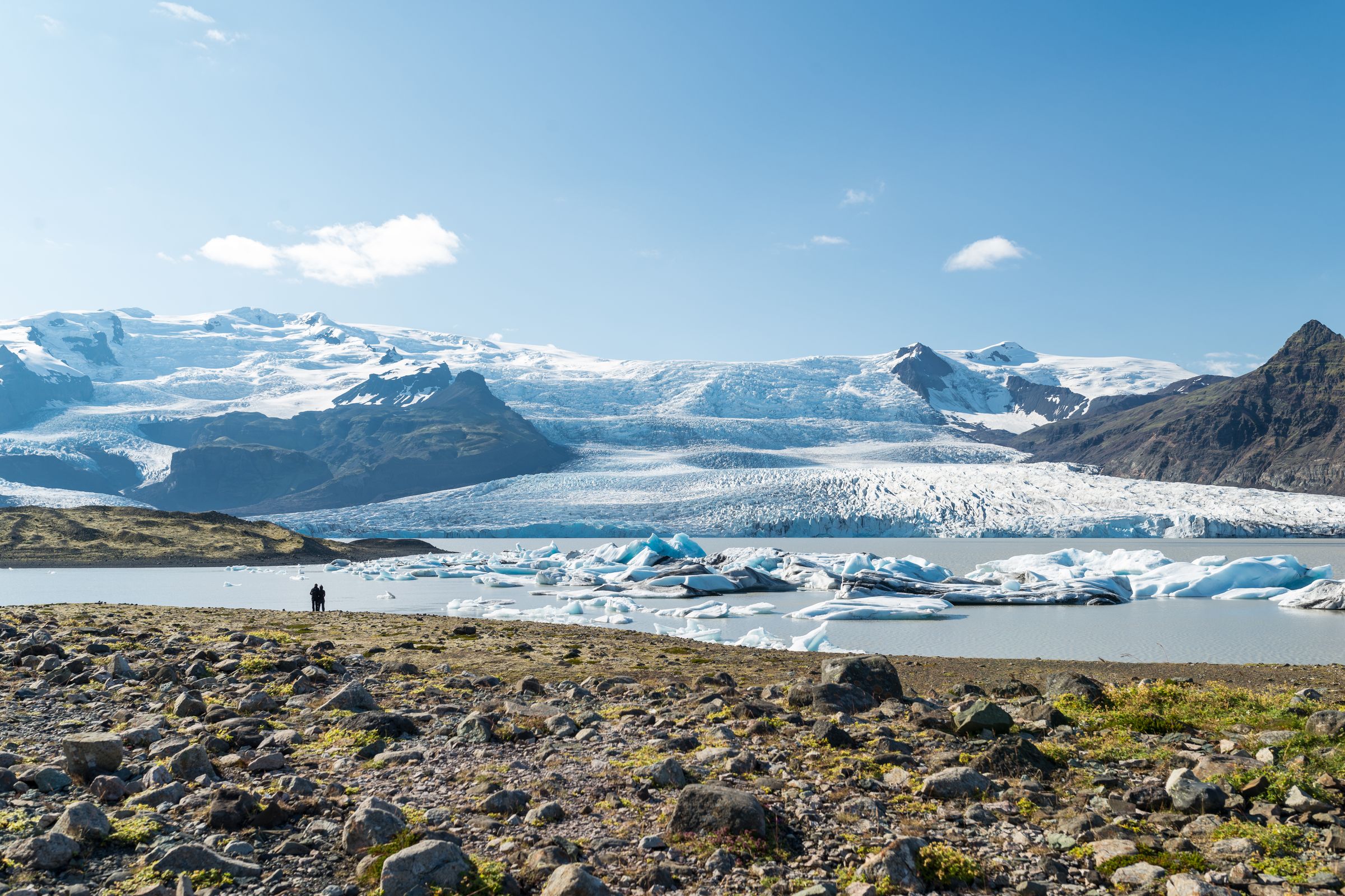 Two people stand on a rocky shore overlooking a glacial lagoon filled with blue icebergs, with a large glacier and mountains in the background under a blue sky.