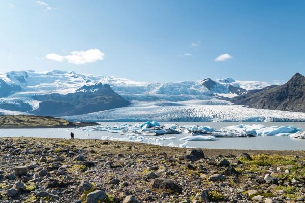Two people stand on a rocky shore overlooking a glacial lagoon filled with blue icebergs, with a large glacier and mountains in the background under a blue sky.