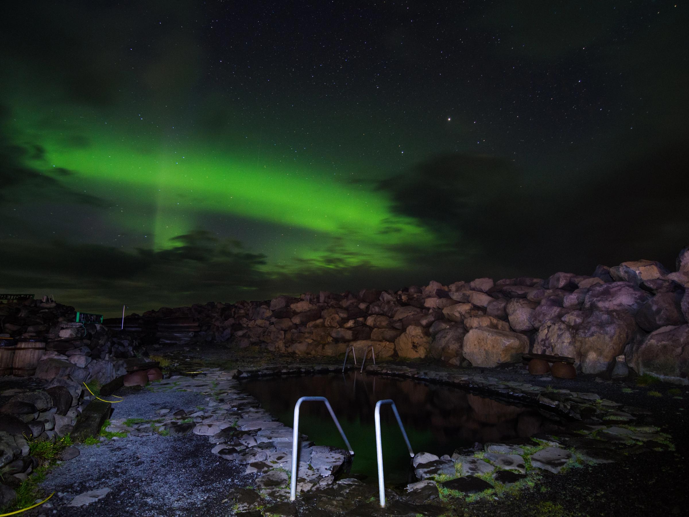 Grettislaug hot spring under the northern lights in North Iceland, surrounded by rugged rocks and night sky.