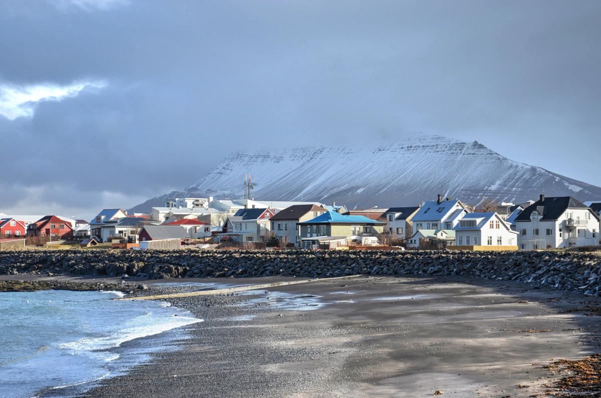 A coastal town with colorful houses by a rocky beach, backed by a snow-capped mountain under a cloudy sky.