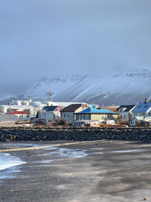A coastal town with colorful houses by a rocky beach, backed by a snow-capped mountain under a cloudy sky.