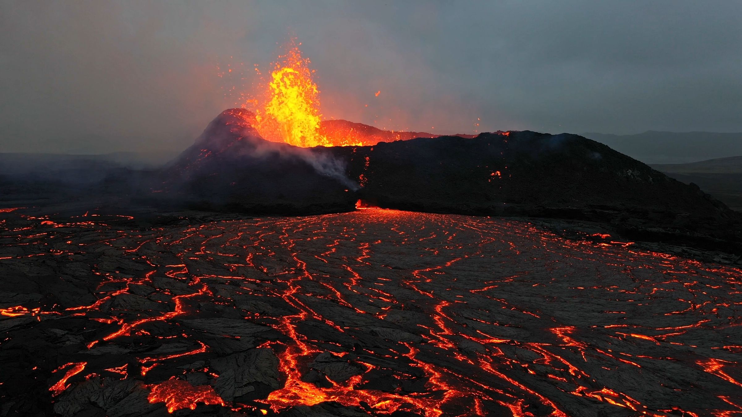 A volcano erupts with a fiery plume, and bright orange lava flows across a dark landscape under a grey sky.
