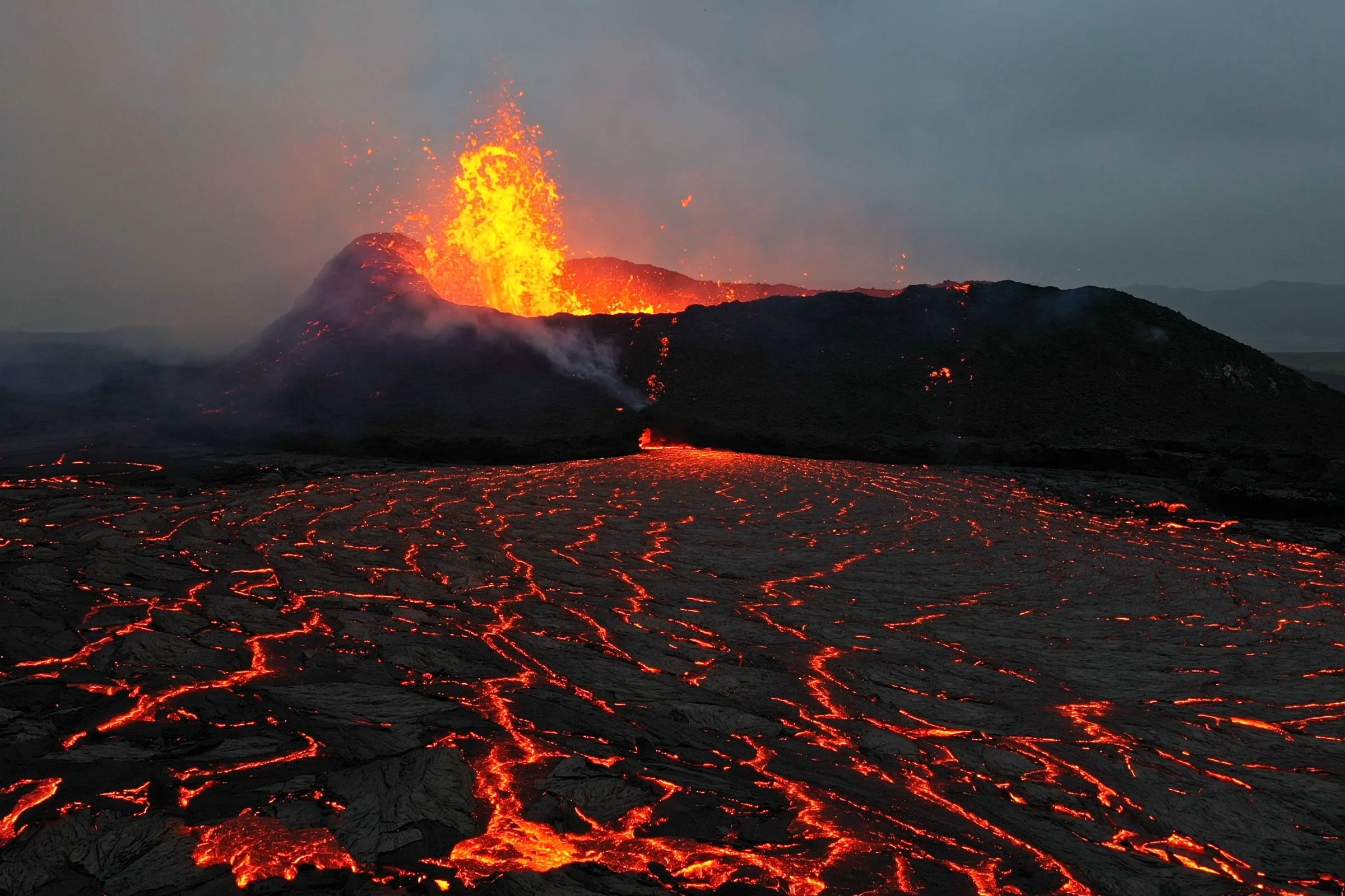 A volcano erupts with a fiery plume, and bright orange lava flows across a dark landscape under a grey sky.