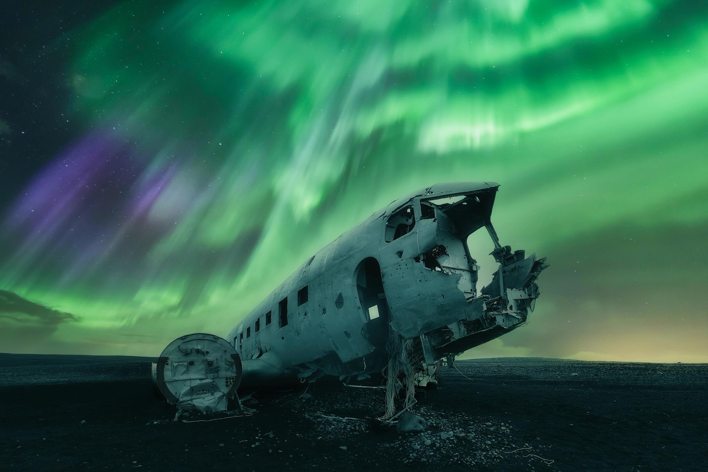 Wrecked plane fuselage on dark ground under vivid green and purple aurora borealis.