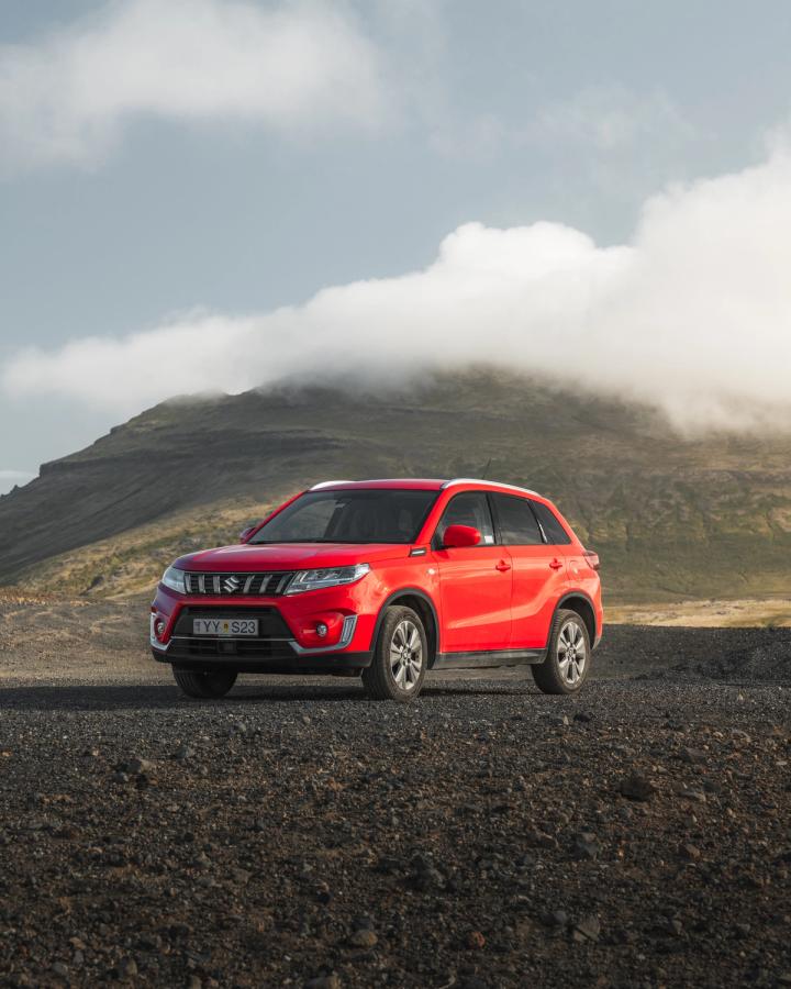 Vue de trois quarts avant d’un SUV Suzuki Vitara garé sur du gravier, avec des nuages bas au-dessus d’une montagne