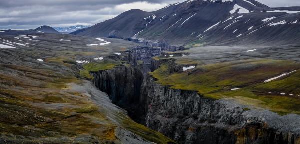 A deep, narrow canyon cuts through a rugged landscape with sparse green and brown vegetation, snow patches, and distant mountains under a cloudy sky.