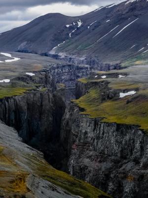 A deep, narrow canyon cuts through a rugged landscape with sparse green and brown vegetation, snow patches, and distant mountains under a cloudy sky.