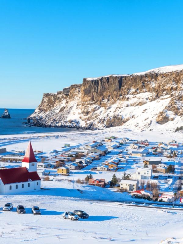 Snow-covered coastal village with a red-roofed church, sea stacks in the ocean, and snow-dusted cliffs under a clear blue sky.
