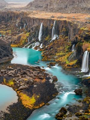 Un río de color turquesa brillante fluye a través de un profundo cañón con numerosas cascadas que caen por acantilados oscuros cubiertos de un follaje otoñal escaso.