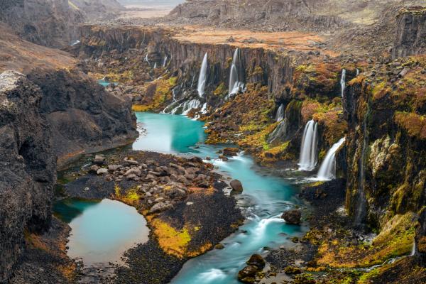 Turquoise river winding through a dark, rocky canyon with multiple waterfalls and patches of colorful vegetation.