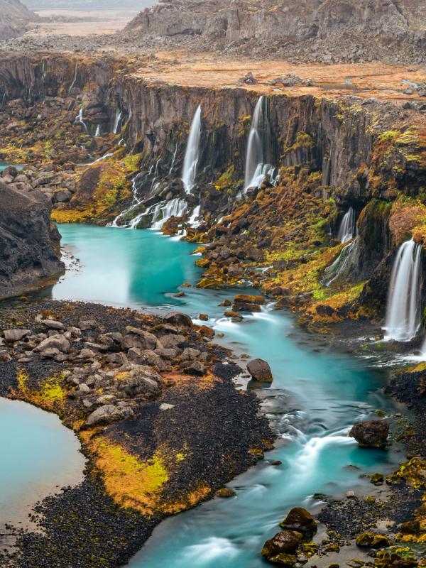 A bright turquoise river flows through a deep canyon with numerous waterfalls cascading down dark cliffs covered in sparse autumn foliage.