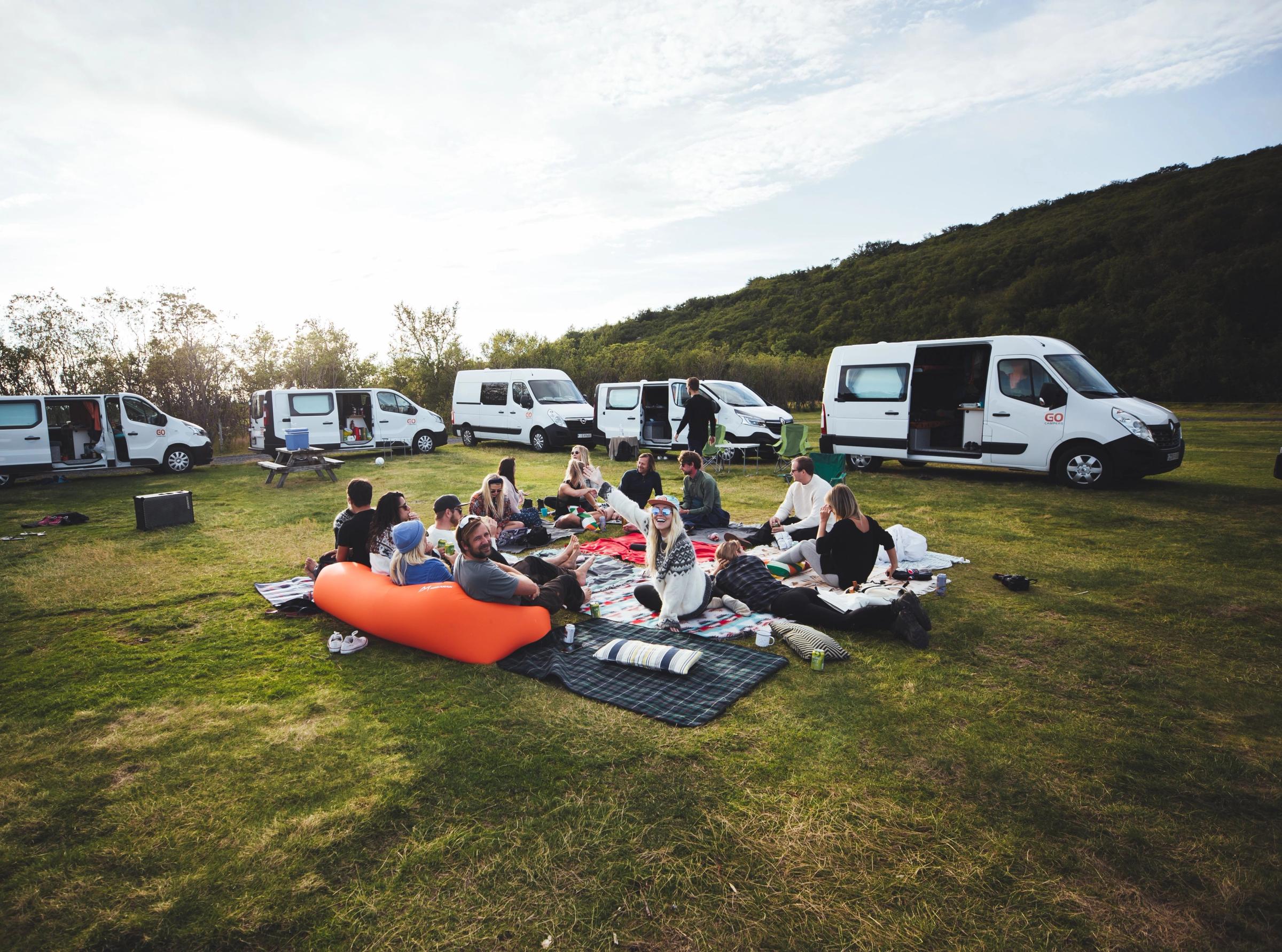 People picnic on blankets in a grassy field surrounded by rental campervans.