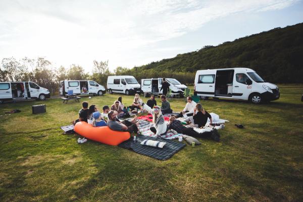Grupo de personas relajándose en un campo cubierto de hierba rodeado de furgonetas camper blancas con las puertas abiertas, una colina boscosa en el fondo.