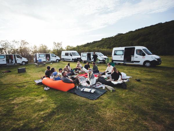 People picnic on blankets in a grassy field surrounded by rental campervans.