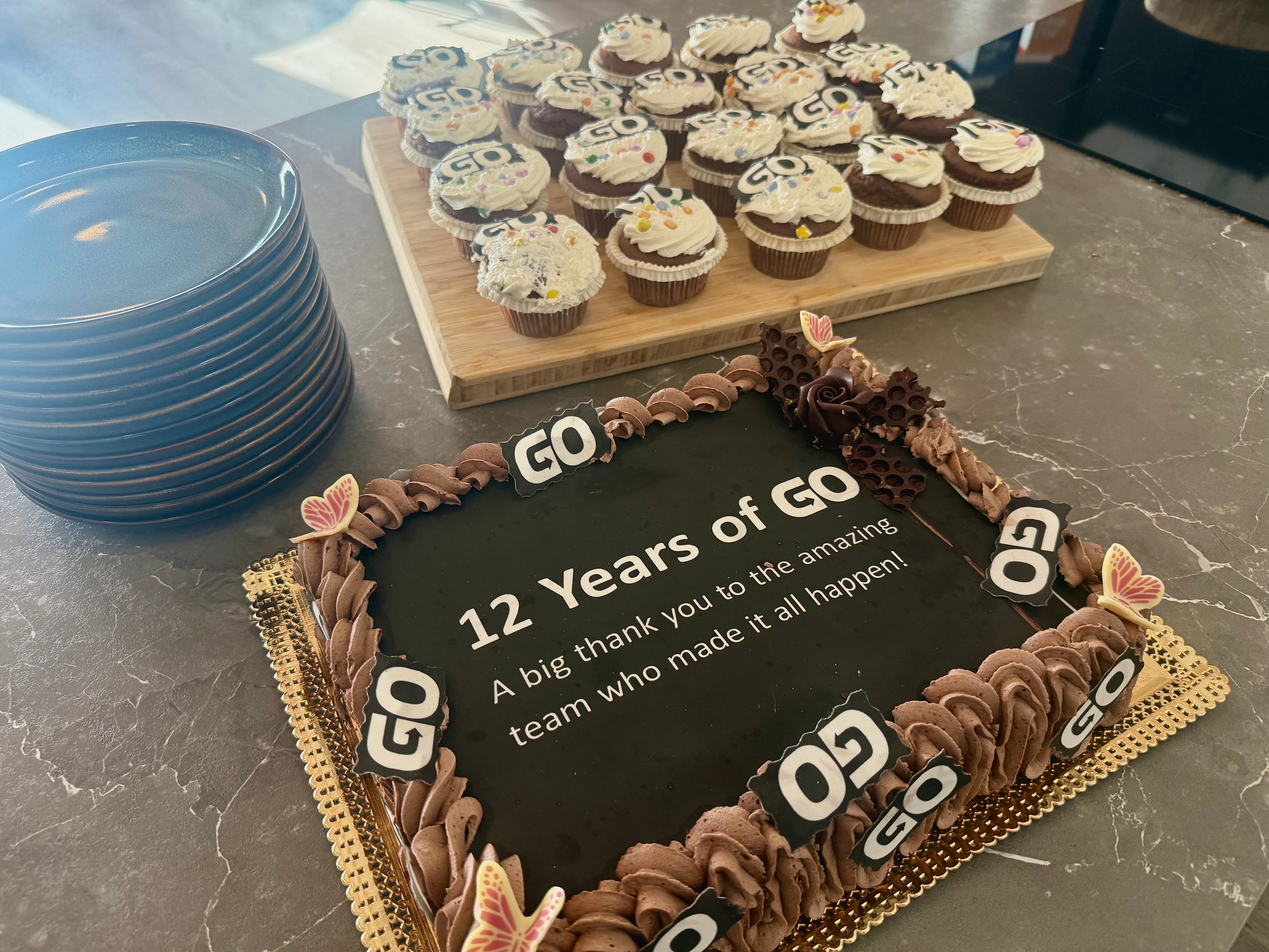 Cake saying "12 Years of GO" with a thank you message, surrounded by "GO" logo cupcakes and a stack of plates.