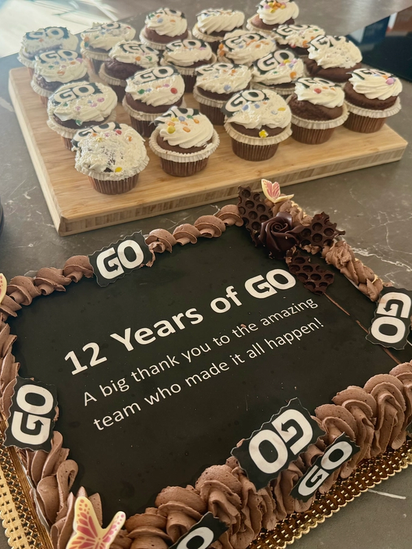 Cake saying "12 Years of GO" with a thank you message, surrounded by "GO" logo cupcakes and a stack of plates.