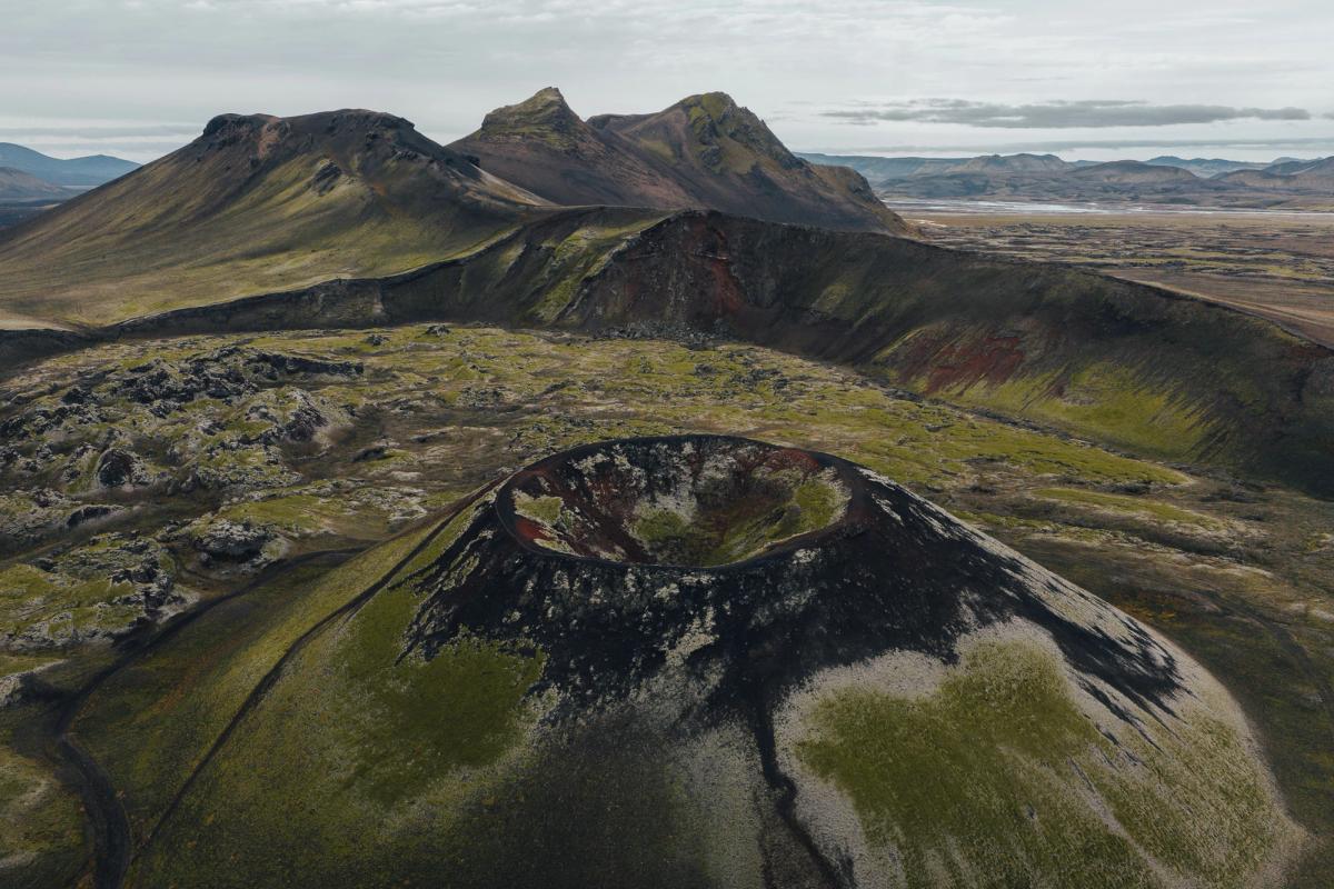 Aerial view of a volcanic landscape featuring a moss-covered crater, dark lava fields, and distant hills under a cloudy sky.