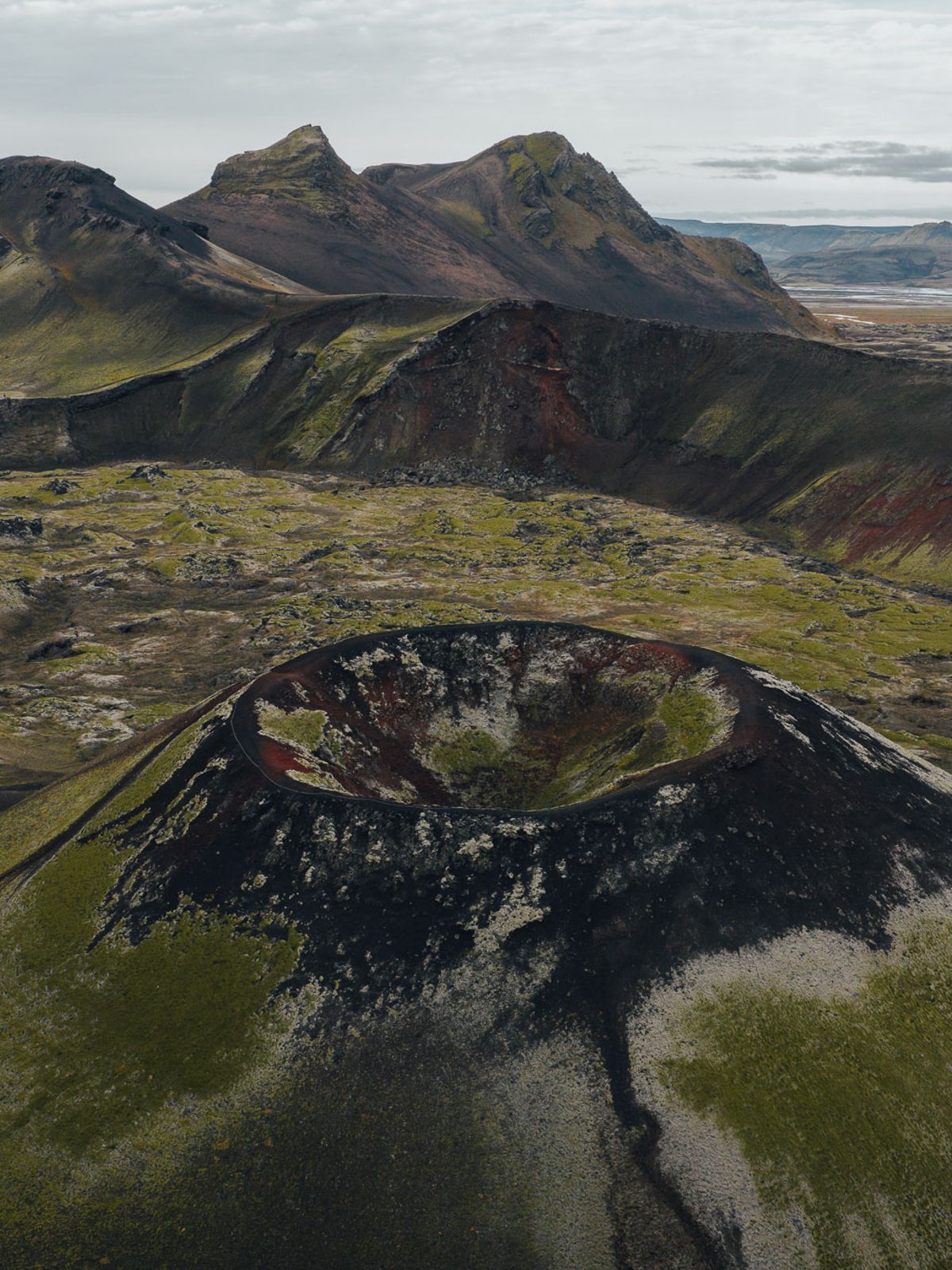 Aerial view of a volcanic landscape featuring a moss-covered crater, dark lava fields, and distant hills under a cloudy sky.