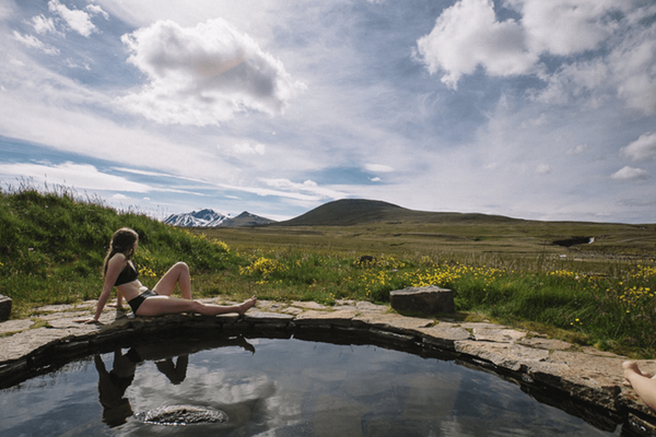 a woman in a bikini sits in a pool with mountains in the background at laugafell in iceland.