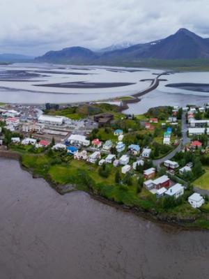 Aerial panoramic of a coastal town with scattered buildings, surrounded by dark water and mudflats, a long causeway, and distant snow-capped mountains under a cloudy sky.