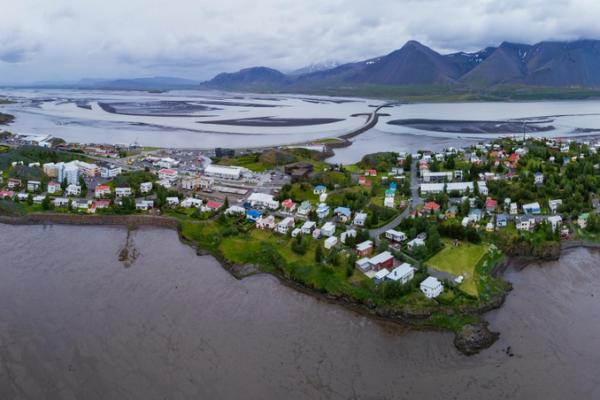 Aerial view of a coastal town on a peninsula, bordering a muddy bay with a causeway, and mountains under an overcast sky.