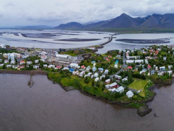 Aerial panoramic view of a coastal town on a peninsula, surrounded by water and mudflats, with mountains under a cloudy sky.