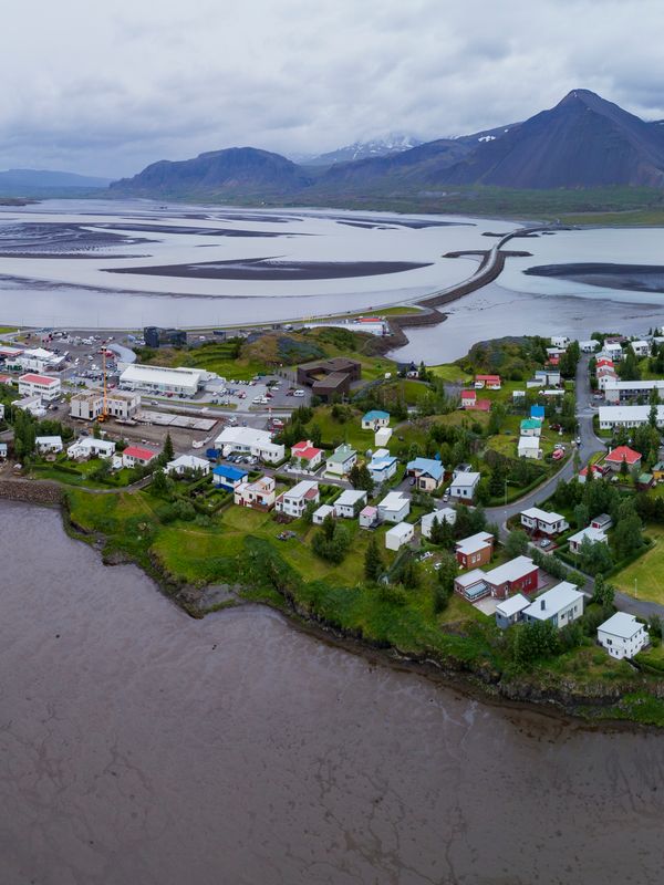 Aerial panoramic of a coastal town with scattered buildings, surrounded by dark water and mudflats, a long causeway, and distant snow-capped mountains under a cloudy sky.