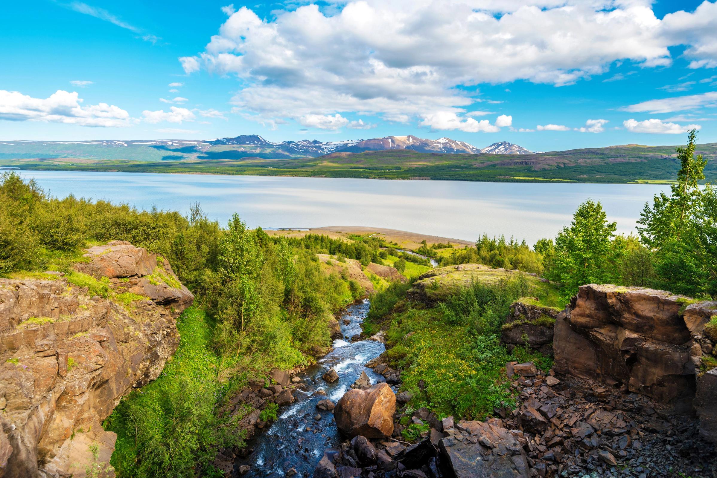 A stream flows through a rocky gorge filled with green trees, leading to a large lake with mountains in the background under a blue, cloudy sky.