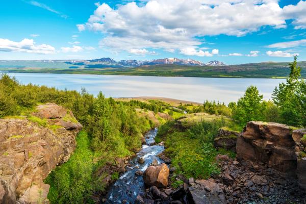A stream flows through a rocky, green landscape towards a large lake and distant mountains under a blue, cloudy sky.