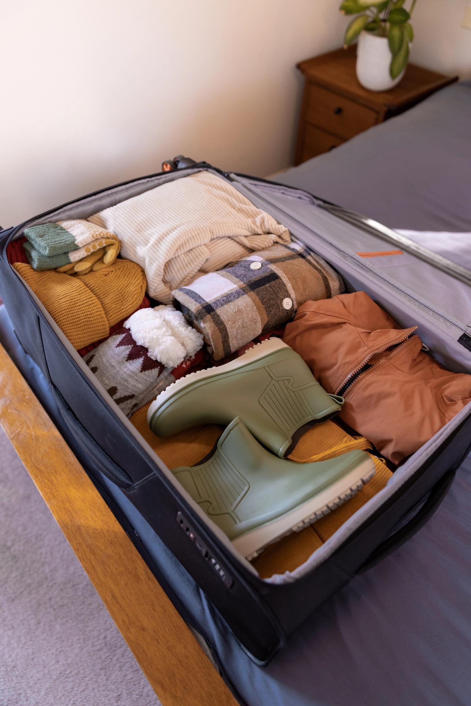 An open suitcase filled with neatly packed warm-toned clothing and sage green rain boots.