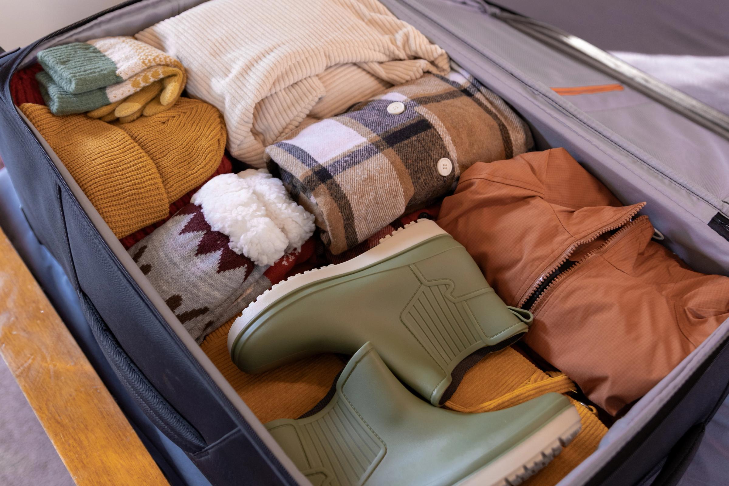 An open suitcase filled with neatly packed warm clothing and a pair of green boots.